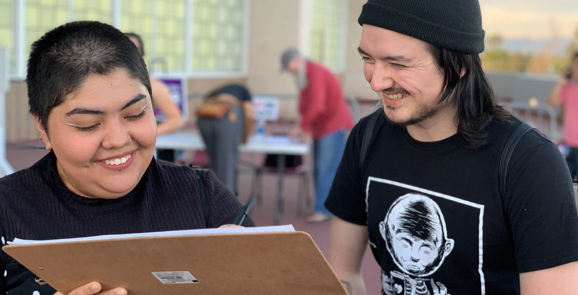 Two young adults smile while filling out a form on a clipboard at an outdoor event