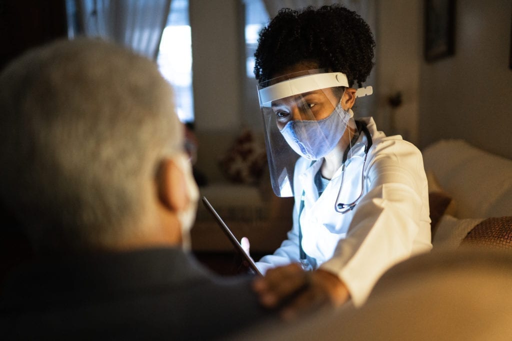 A doctor wearing a face covering and face shield visits a patient's home and listens to the patient while holding their shoulder