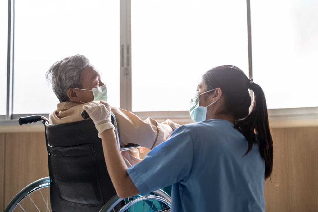 Asian nurse taking care of mature male patient sitting on wheelchair in hospital. Young woman and old man wearing surgical face mask for protection of covid 19 pandemic. Girl smile to elderly man. A patient in a wheelchair shares a laugh with a medical staff member